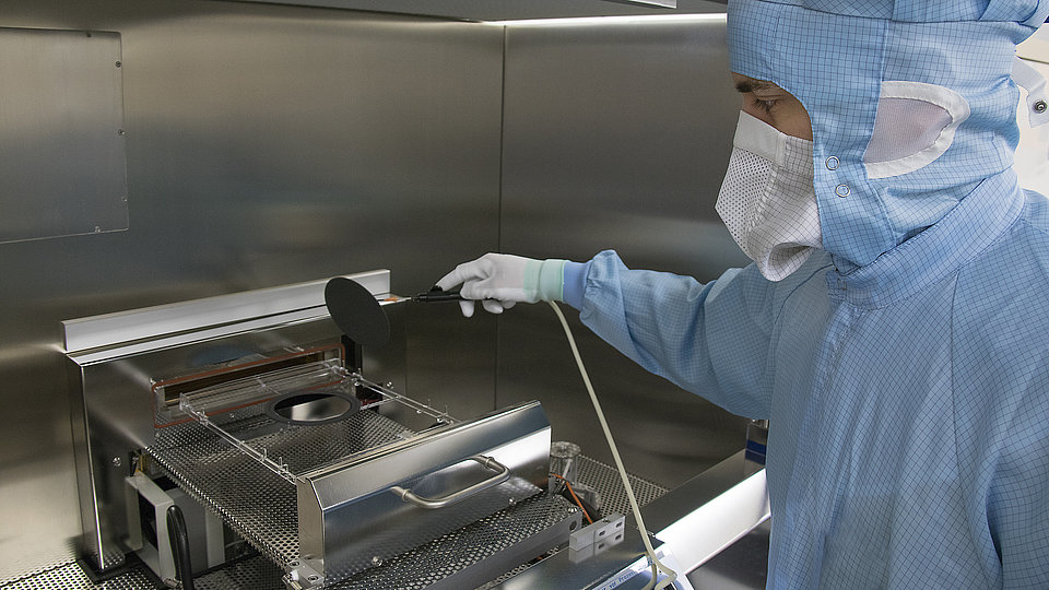 A person in protective clothing, including a face mask and hood, is working in a cleanroom environment. They are using a tool to inspect or manipulate equipment inside a metallic workstation.
