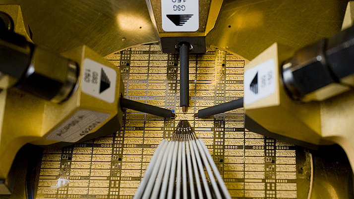 Close-up of a semiconductor wafer being processed in a machine. Metallic pins are positioned above the wafer, which has a grid-like pattern.