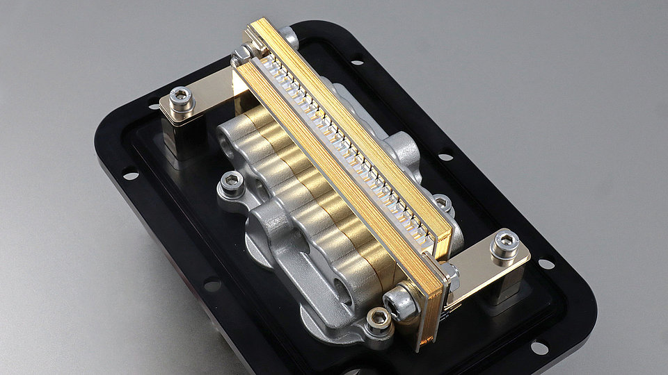 Laser stack made of gray metal and gold-colored connections. It is mounted on a black plate and has several screws and brackets. 