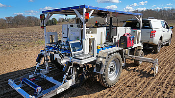 An agricultural vehicle with a sensor system is standing on a trailer in a field. It has large tires, a platform with a laptop and various technical components. A pickup truck can be seen in the background pulling the trailer. The sky is clear with some clouds. 